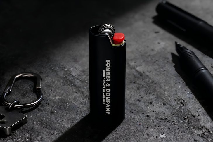 Black lighter with 'Bomber & Company' branding on a dark surface with a pen and keychain.